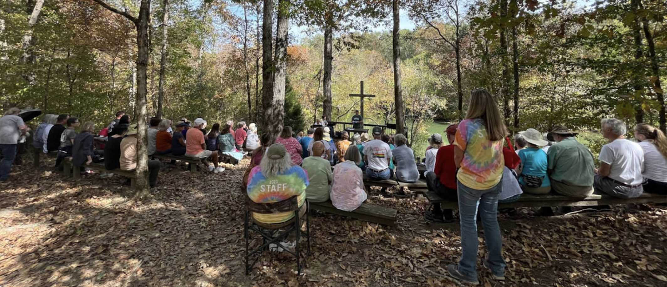 A group of people sit on chairs in a wooded clearing, facing a man speaking on an open stage in front of a cross.