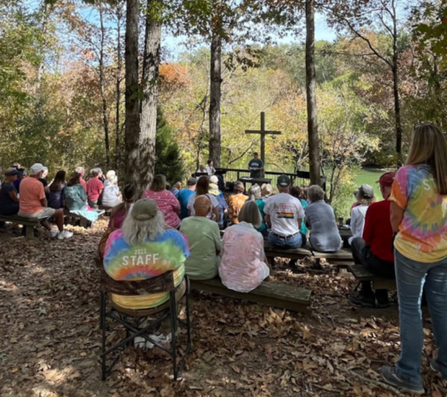 A group of people sit on chairs in a wooded clearing, facing a man speaking on an open stage in front of a cross.