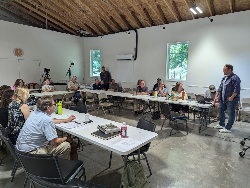 Around 12-15 people sit at a U-shaped series of fold-out tables, looking at a presentation that is out of frame. Most people have notebooks or laptops, and a camera is recording the proceedings.