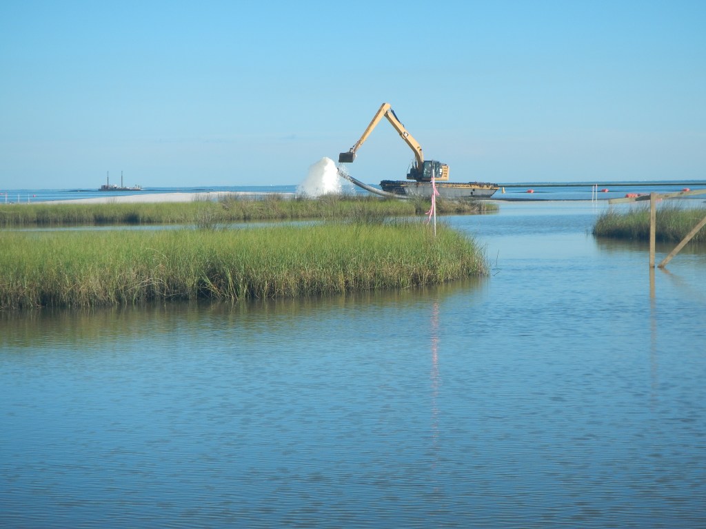 A crane dumps a load of sand onto a beach. The crane is positioned at the edge of the water, and several other grassy inlets into the water are visible.