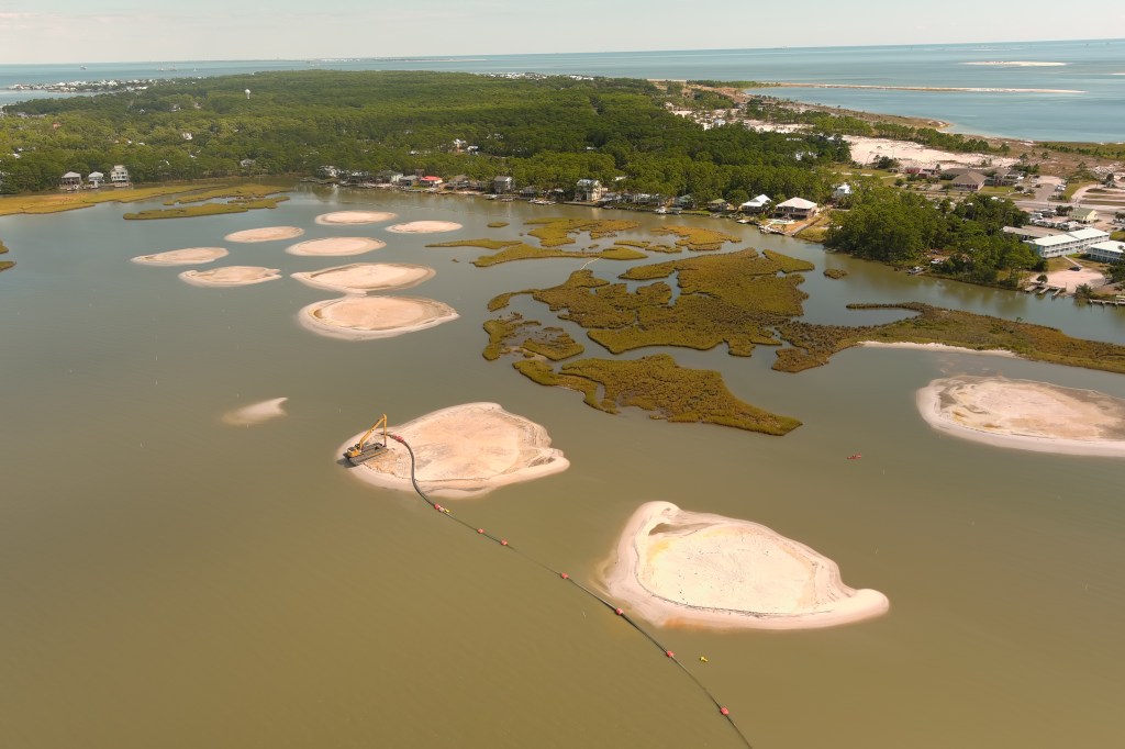 An aerial view of several sand mounds in front of existing marshland and a row of coastal homes. The forested east end of the island is visible in the background, with the gulf on the other side. A crane is at work on one of the sand mounds.