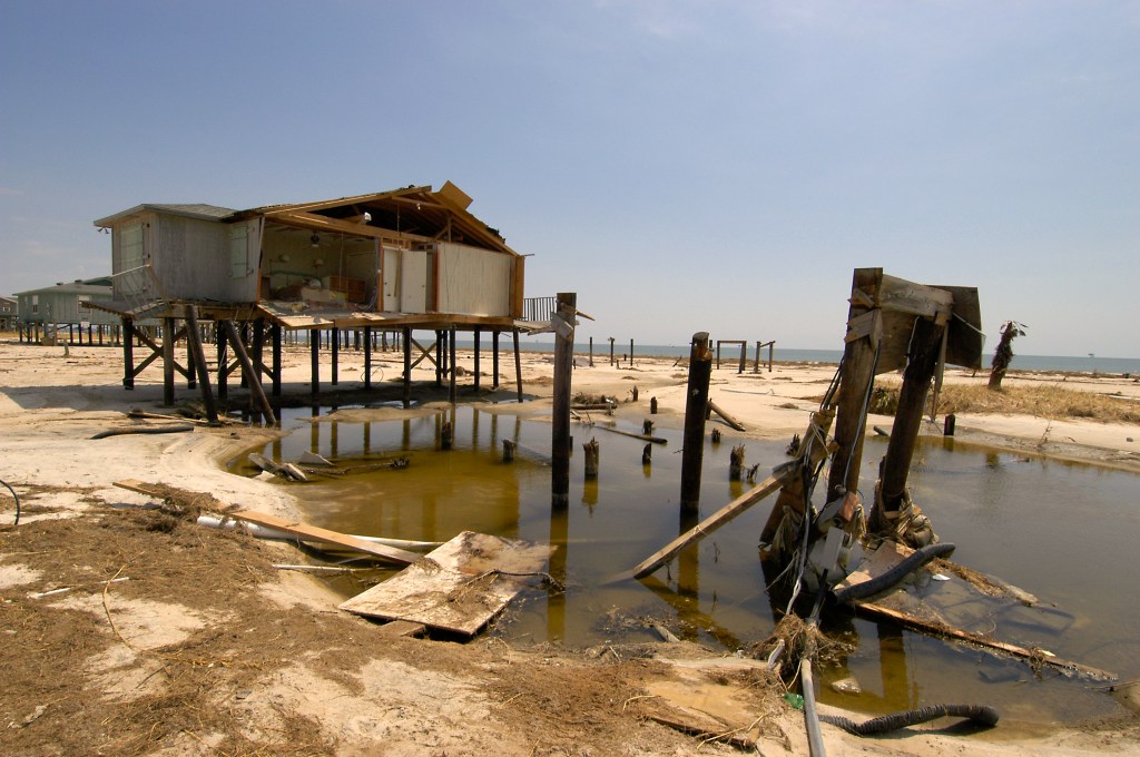 An elevated home has been sheared in half, with a bedroom now visible from the outside. Other pilings to elevate other structures are snapped, toppled or otherwise damaged and surrounded by debris and standing water.