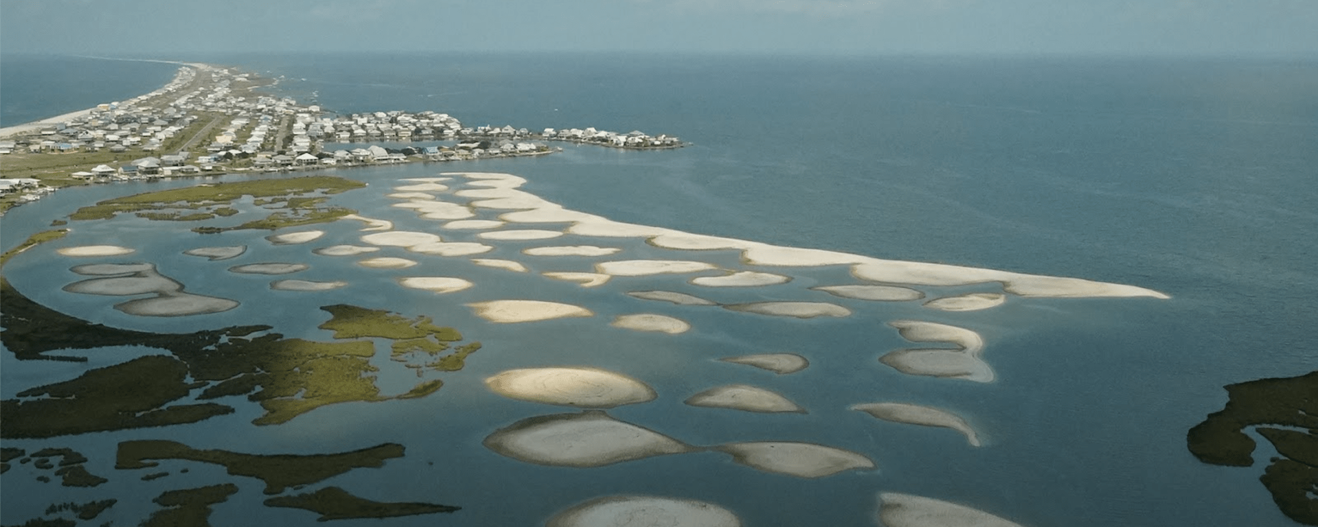 An aerial view of a curved coastline with sand mounds dotted through the water. Homes are visible on the spit of land in the background.