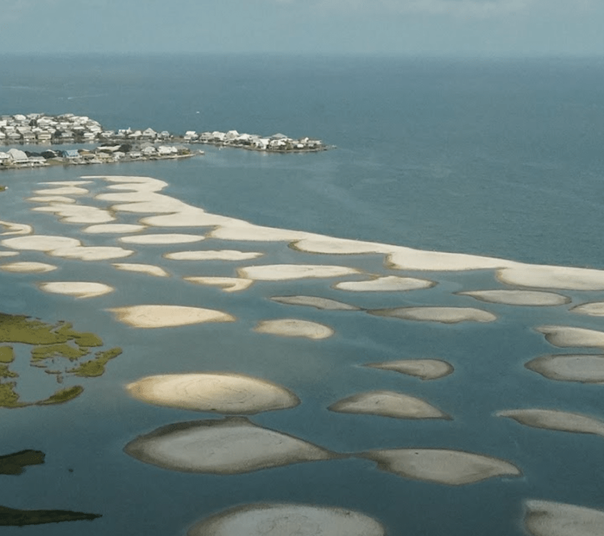 An aerial view of a curved coastline with sand mounds dotted through the water. Homes are visible on the spit of land in the background.
